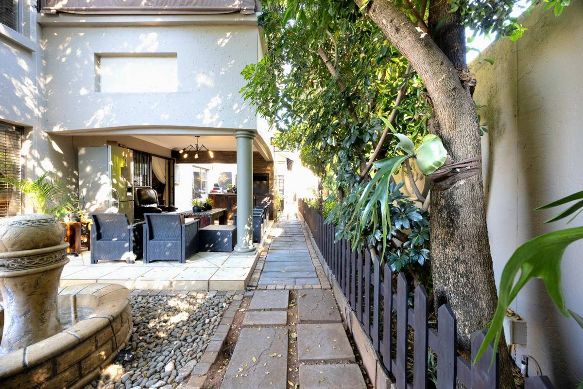 A well-maintained rear garden area featuring a stone-paved walkway, a covered patio space, and a decorative stone fountain alongside a picket fence.