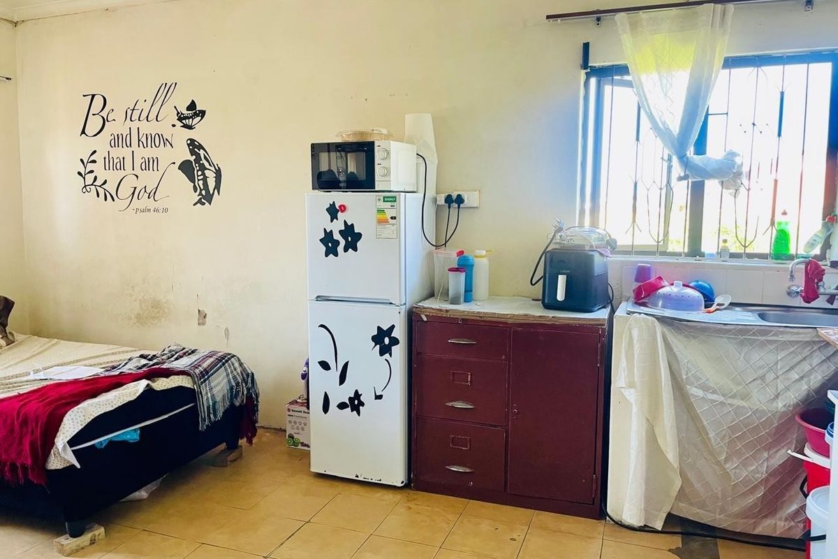 A basic kitchen area featuring tiled flooring, a stainless steel sink, and security bars on the window, currently showing signs of wear and maintenance needs.