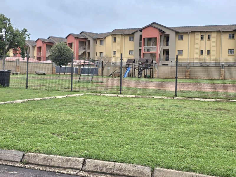 A communal outdoor area featuring a lawn and playground equipment, situated behind a residential apartment complex with secure perimeter fencing.