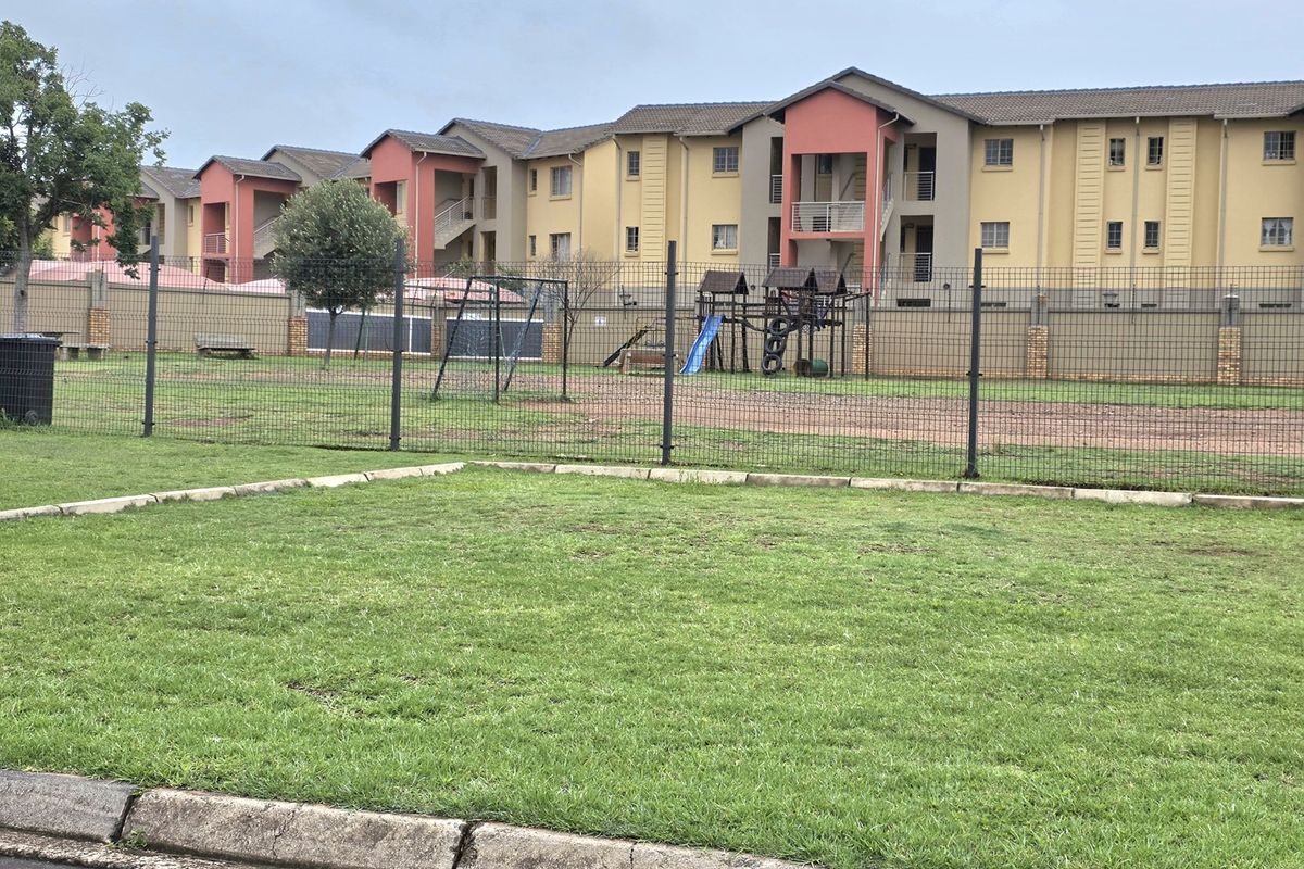 A communal outdoor area featuring a lawn and playground equipment, situated behind a residential apartment complex with secure perimeter fencing.