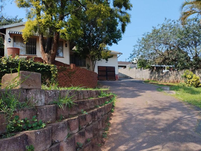 Front exterior view of the property featuring a paved driveway, brick retaining wall, and garage access with established trees and boundary walling.