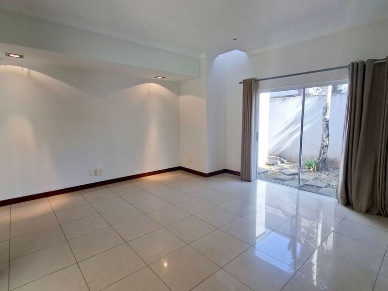 A bright, empty living room featuring large format floor tiles, recessed lighting, a skylight, and sliding glass doors leading to a private outdoor area.