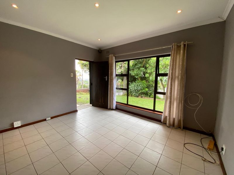 A bright, empty living room featuring neutral tiled flooring, recessed lighting, and large windows providing natural light and garden views.
