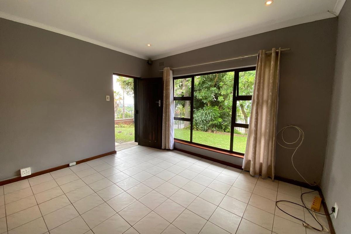 A bright, empty living room featuring neutral tiled flooring, recessed lighting, and large windows providing natural light and garden views.