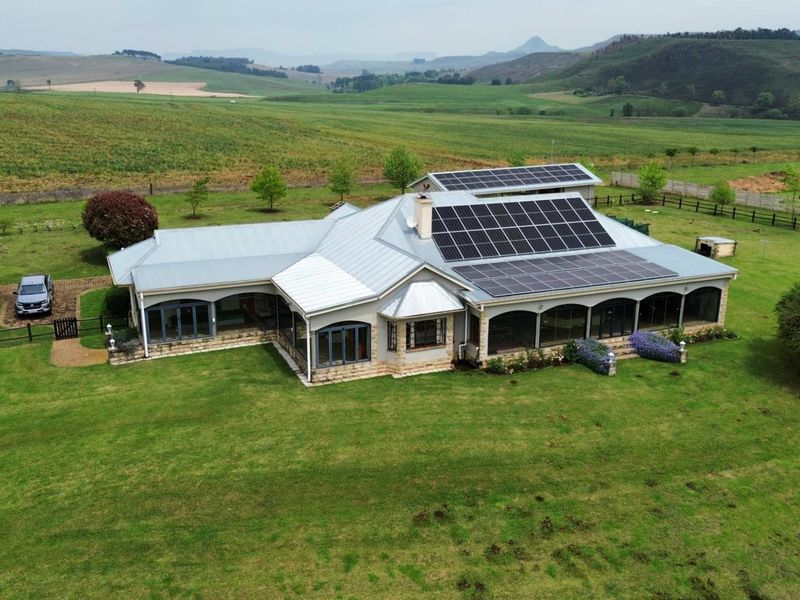 Aerial view of a well-maintained farmhouse featuring a metal roof, extensive solar panel installation, and a spacious covered patio set in a rural landscape.