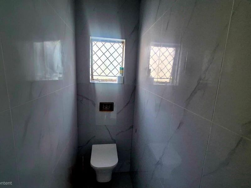 A compact, modern bathroom featuring marble-effect wall tiles, a wall-mounted toilet with a flush plate, and a window with a security grille.