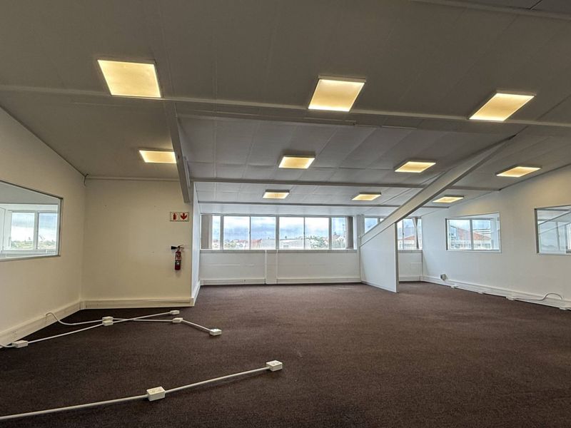 A spacious, open-plan office area featuring neutral walls, carpeted flooring, recessed ceiling lights, and large windows providing ample natural light.