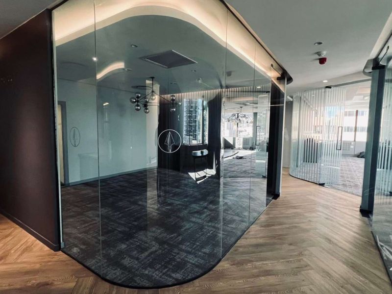 Modern office space featuring curved glass partitioning, recessed ceiling lighting, and a combination of herringbone wood and carpeted flooring.