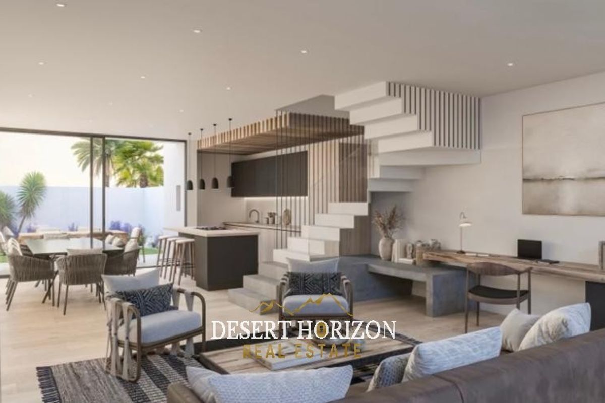Modern open-plan living area featuring a sleek kitchen, integrated desk space, and a striking staircase, flowing seamlessly to an outdoor patio through sliding doors.