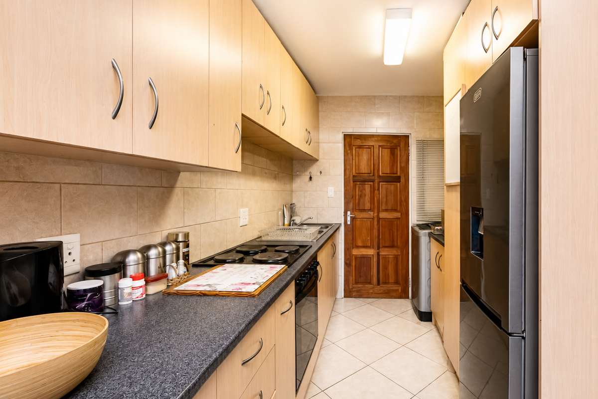 A functional galley-style kitchen featuring light wood cabinetry, dark laminate countertops, tiled backsplash, and built-in electric cooking appliances.