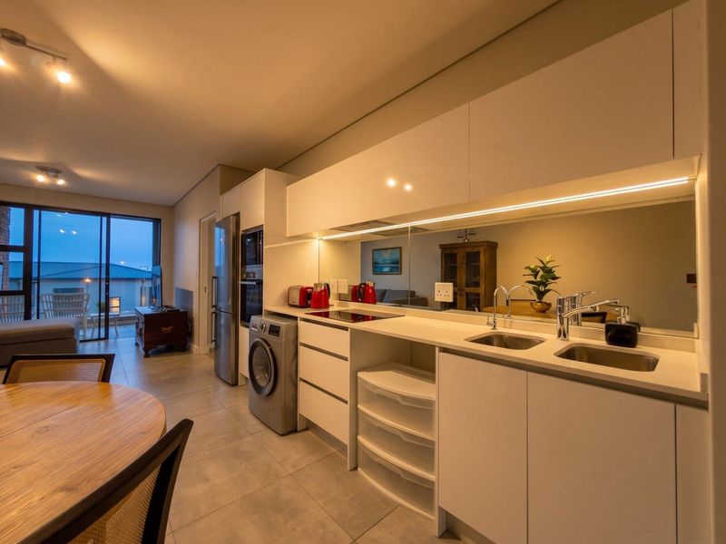 Modern open-plan kitchen featuring sleek white cabinetry, stone countertops, a mirrored splashback, and integrated appliances, flowing seamlessly into the living area.