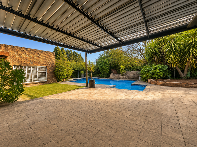 Spacious outdoor entertainment area featuring a covered patio, paved terrace, and a swimming pool set within a landscaped garden.