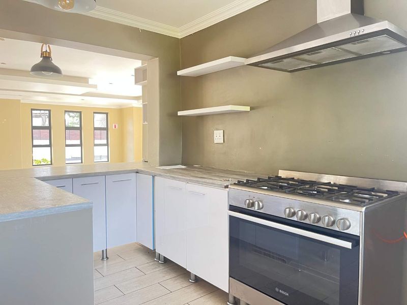 A clean, modern kitchen featuring white cabinetry, laminate countertops, a stainless steel gas stove with extractor fan, and open shelving, flowing into an adjacent living area.