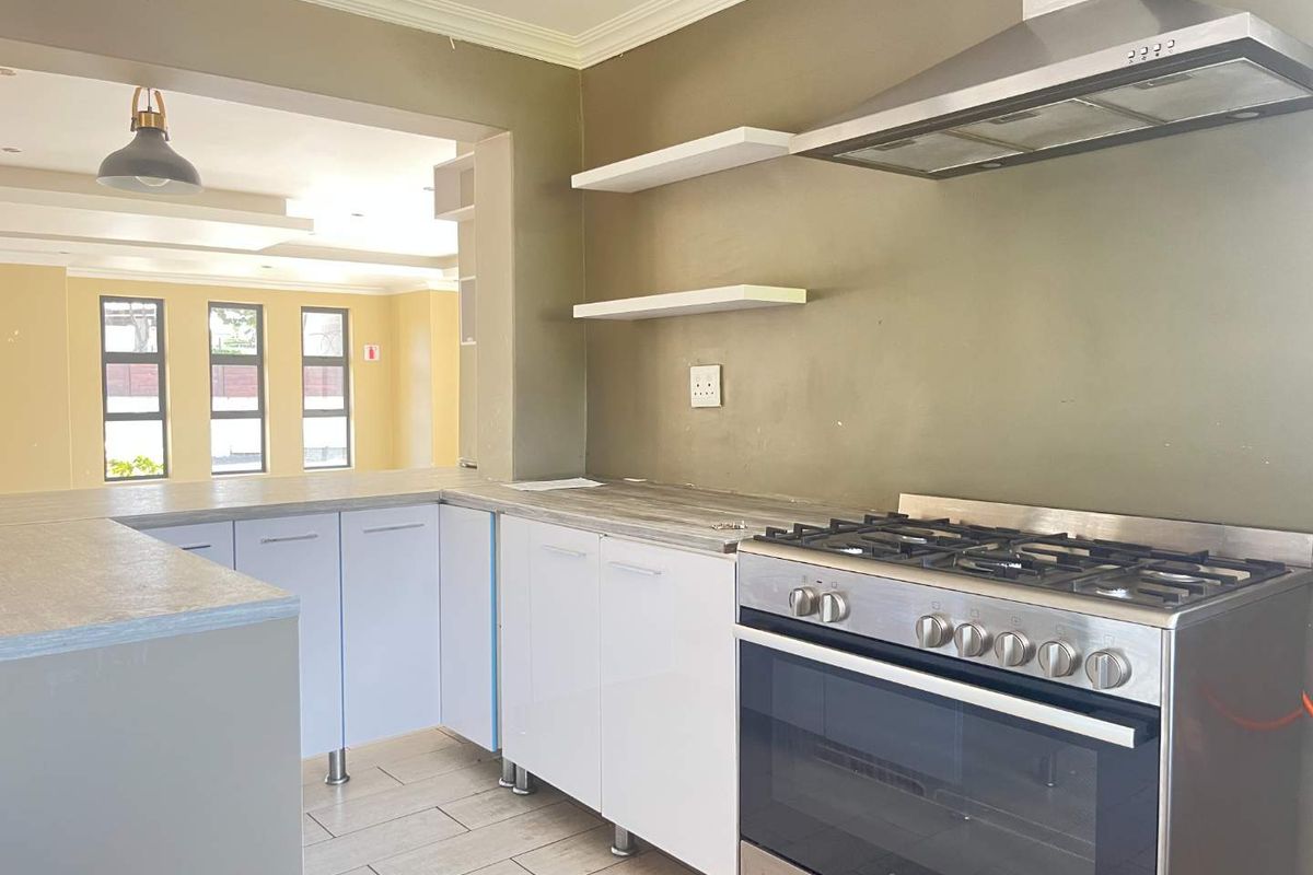 A clean, modern kitchen featuring white cabinetry, laminate countertops, a stainless steel gas stove with extractor fan, and open shelving, flowing into an adjacent living area.