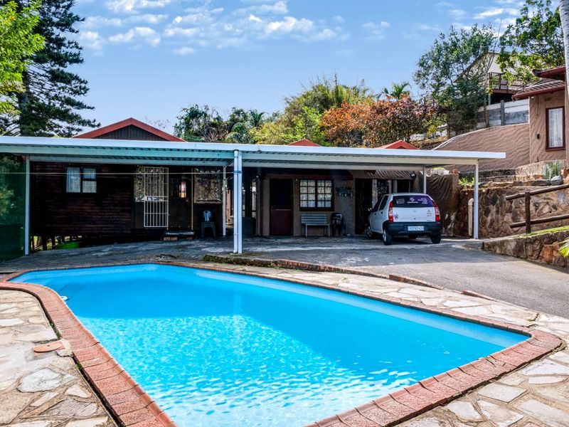 Outdoor pool area featuring a paved deck, stone retaining wall, and a covered carport structure adjacent to the main dwelling.