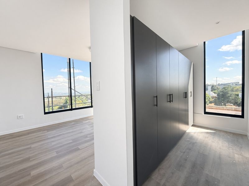 A bright, modern bedroom featuring sleek dark built-in cupboards, light laminate flooring, and expansive windows offering elevated views.