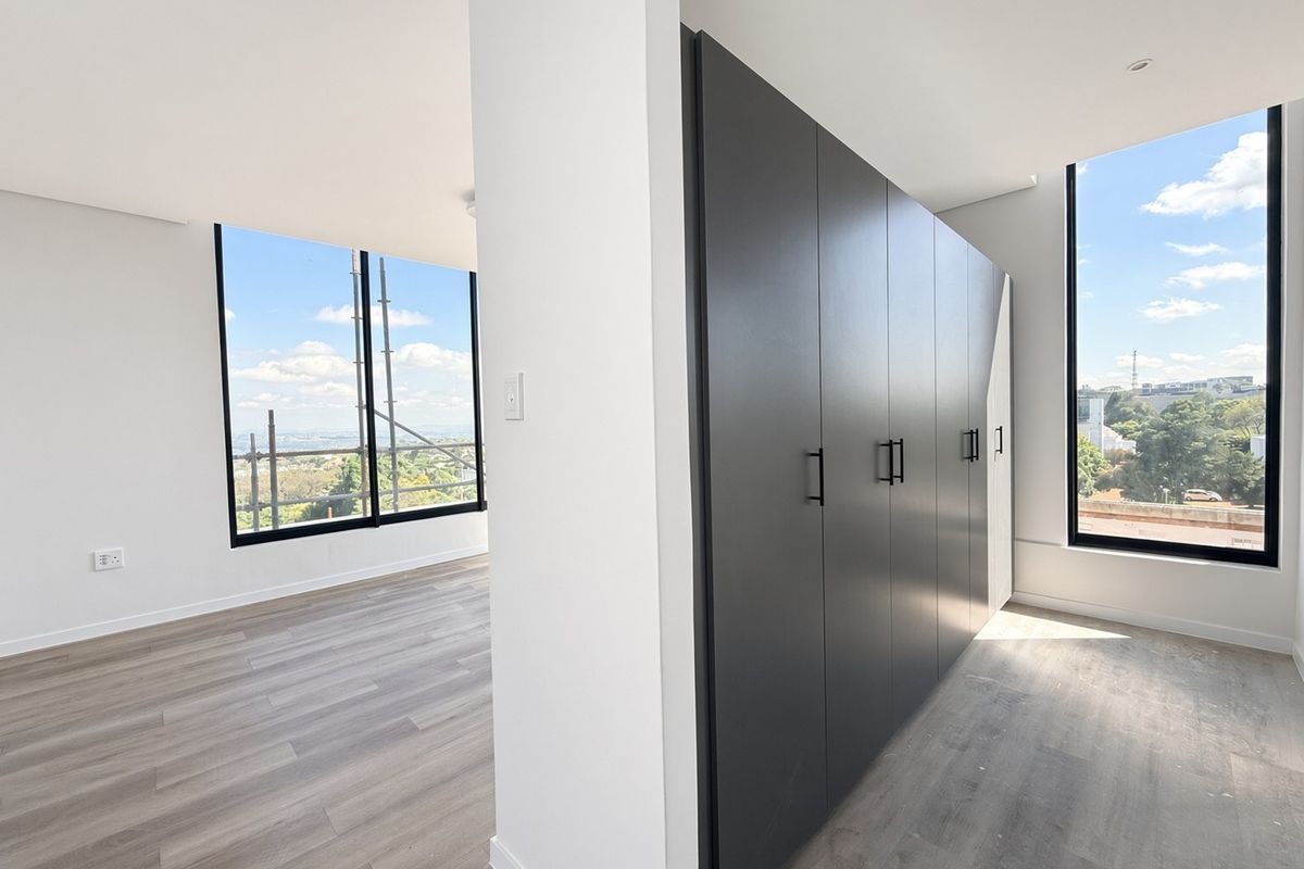 A bright, modern bedroom featuring sleek dark built-in cupboards, light laminate flooring, and expansive windows offering elevated views.