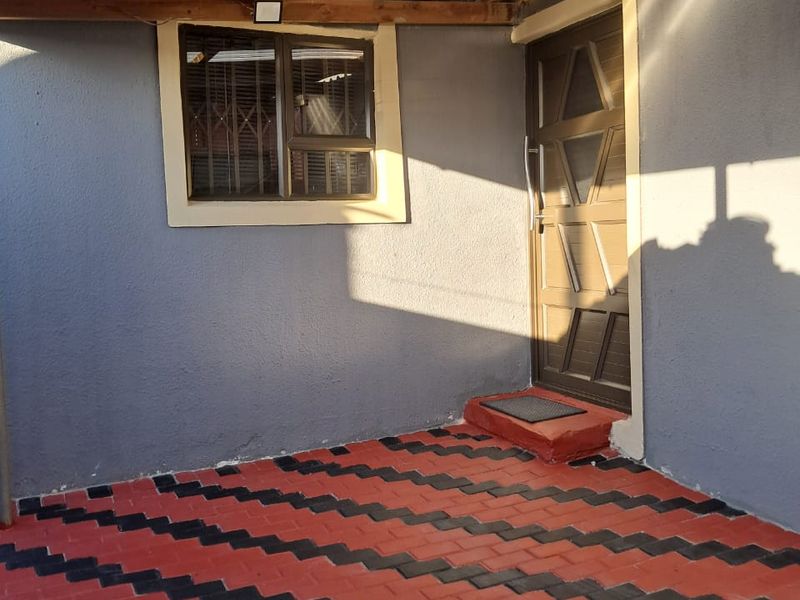 A covered outdoor patio area featuring patterned brick paving, a secure entrance door, and a timber-framed roof structure.