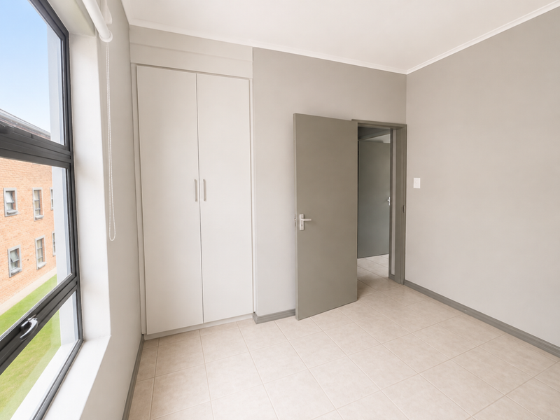 A bright, empty bedroom featuring neutral tiled flooring, built-in cupboards, and a large window providing natural light.
