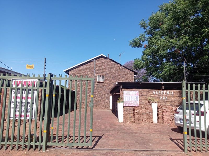 Front view of a brick apartment complex entrance featuring a paved driveway, secure metal gate, electric fencing, and a covered carport area.
