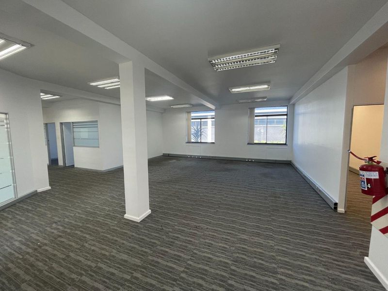 A spacious, open-plan office area featuring neutral carpeted flooring, bright fluorescent lighting, and large windows providing natural light.