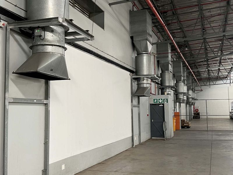 Spacious industrial warehouse interior featuring high ceilings with exposed steel trusses, concrete flooring, and a series of wall-mounted industrial ventilation units.
