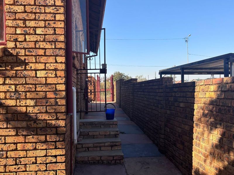 A side exterior view of the house featuring face brick walls, a paved walkway, concrete steps, and a perimeter boundary wall.