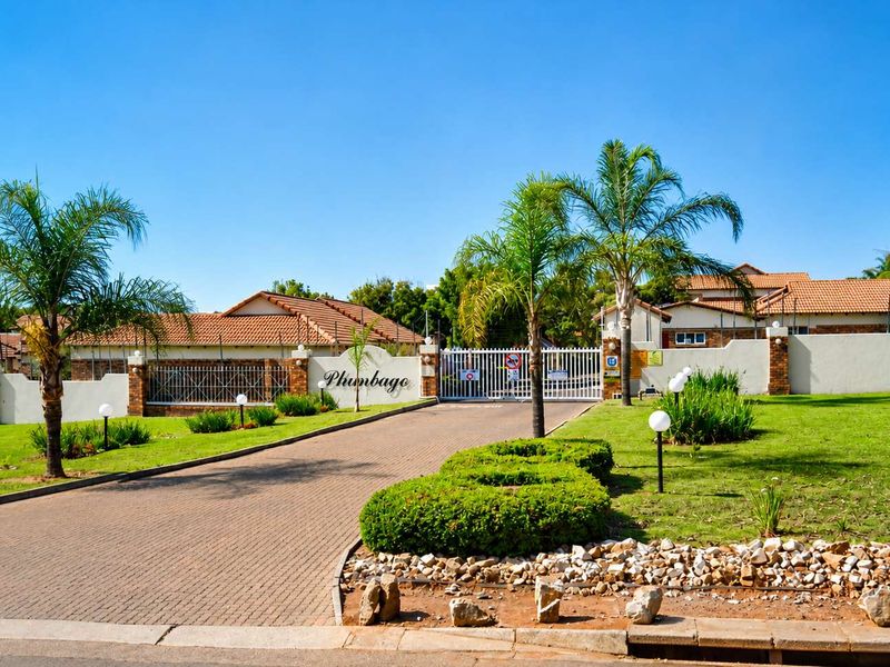 Front entrance view of the Plumbago townhouse complex, featuring a paved driveway, secure gated access, perimeter walling, and well-maintained landscaped gardens.