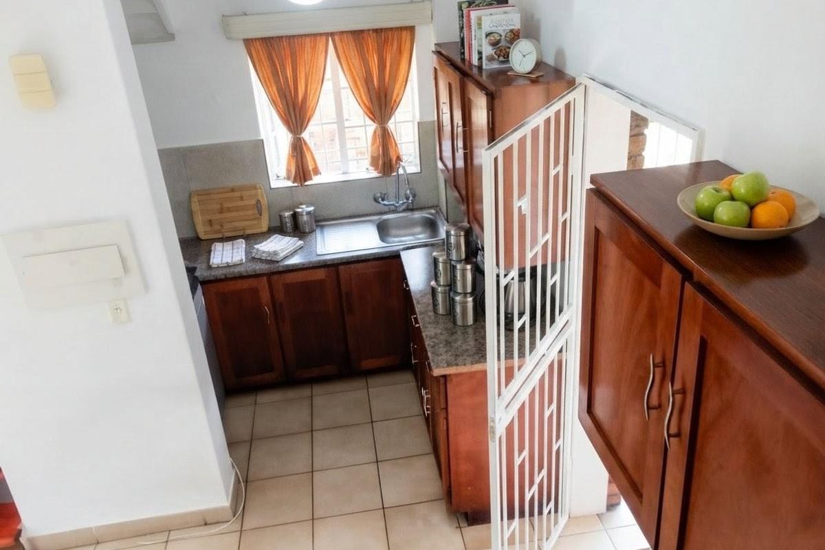 A compact kitchen featuring wooden cabinetry, laminate countertops, a stainless steel sink, and tiled flooring, with a security gate at the entrance.
