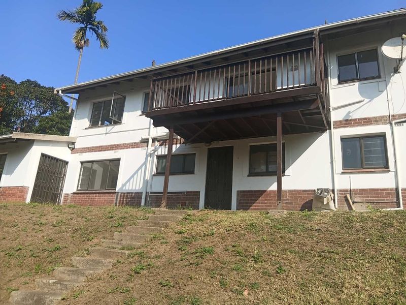 Rear view of a two-story house featuring a raised wooden balcony, brick and plaster exterior, and concrete stairs leading to the garden.