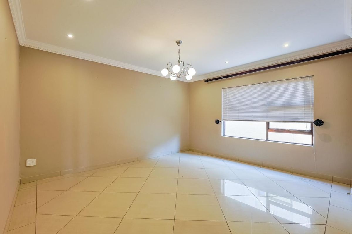 A bright, empty living room featuring neutral tiled flooring, recessed downlighting, decorative ceiling cornicing, and a large window with fitted blinds.