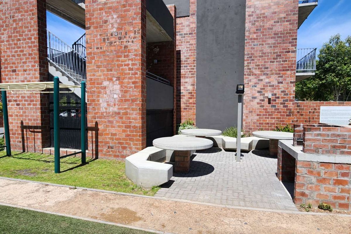 Communal outdoor area featuring a built-in brick braai, concrete seating, and a small fitness frame set against a modern brick apartment building facade.