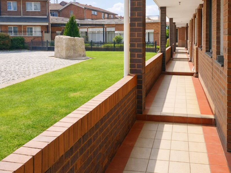 A covered outdoor walkway featuring brick columns and tiled flooring, overlooking a landscaped courtyard area.