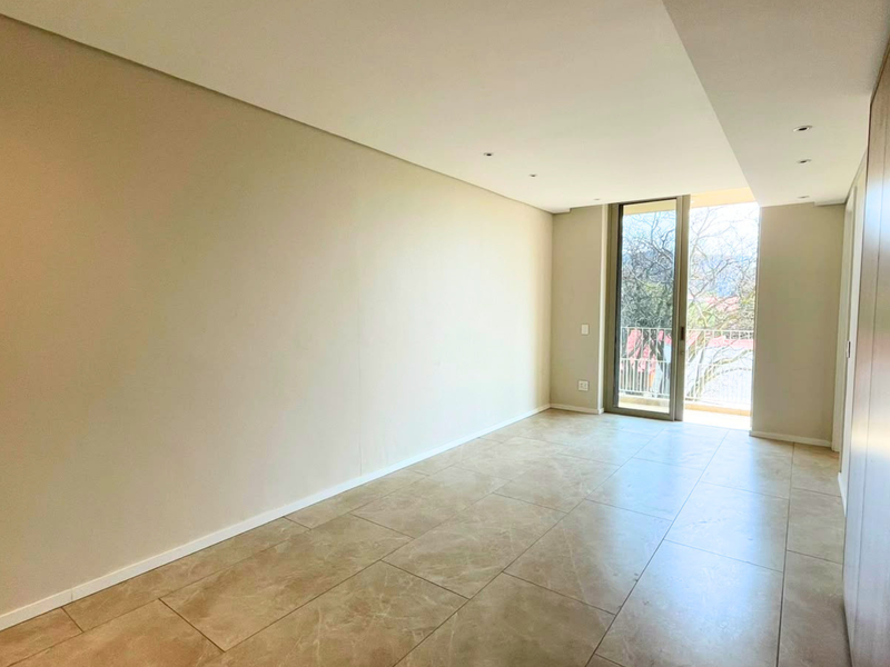 A bright, empty bedroom featuring neutral tiled flooring, recessed lighting, built-in wardrobes, and a sliding glass door leading to a balcony.