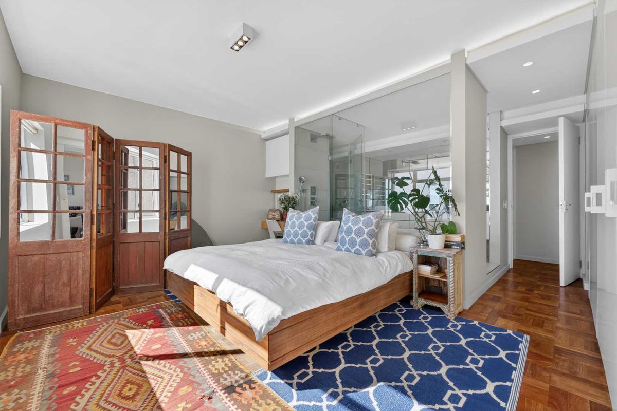 Modern bedroom featuring warm parquet flooring, a sleek glass-enclosed ensuite bathroom, and clean, neutral wall finishes with integrated lighting.