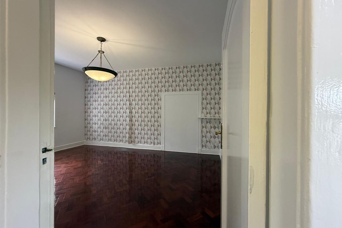 A spacious bedroom featuring dark herringbone parquet flooring, patterned wallpaper, and a central pendant light fixture.