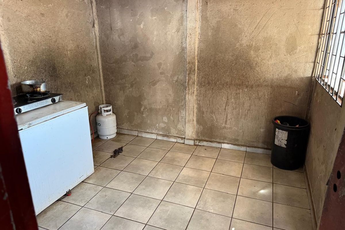 A basic, unfinished kitchen area featuring tiled flooring, security window bars, and a gas hob setup, requiring significant cosmetic renovation.