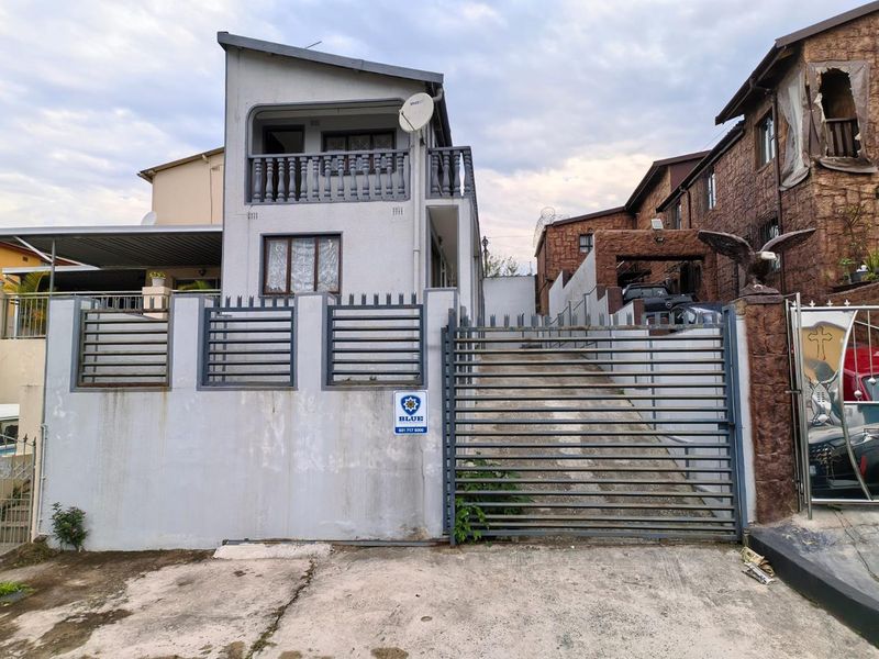 Front elevation of a two-story house featuring a concrete driveway, secure metal gate, and an upper-level balcony with balustrade.