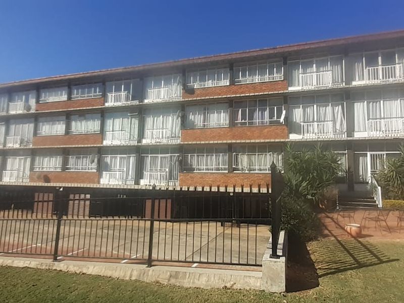 A multi-story apartment building featuring a classic brick facade, private balconies, and a paved parking area with secure perimeter fencing.