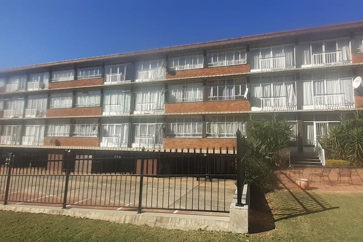 A multi-story apartment building featuring a classic brick facade, private balconies, and a paved parking area with secure perimeter fencing.