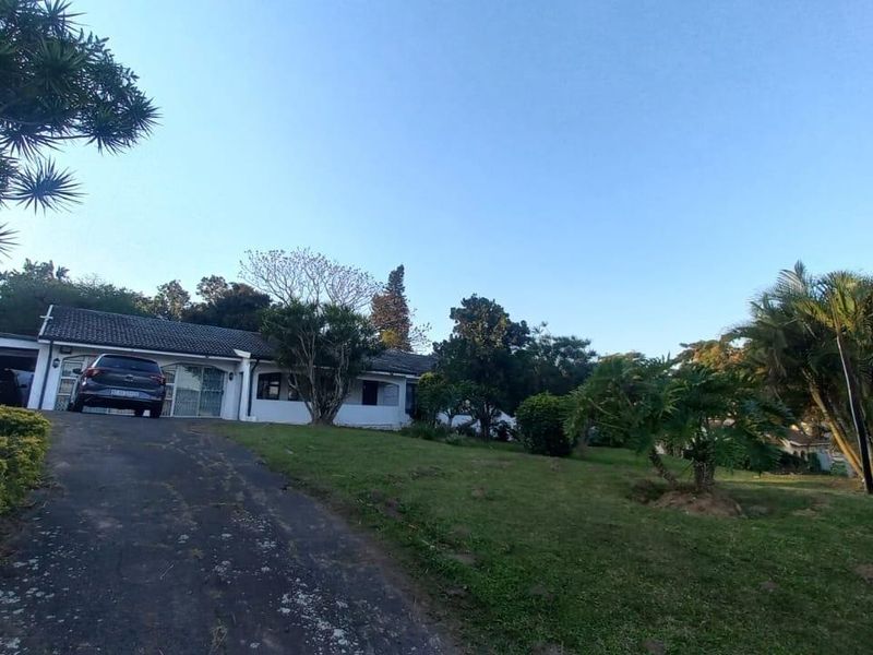 Front exterior view of a single-story house featuring a tiled roof, attached garage, and a long asphalt driveway set within a spacious garden.