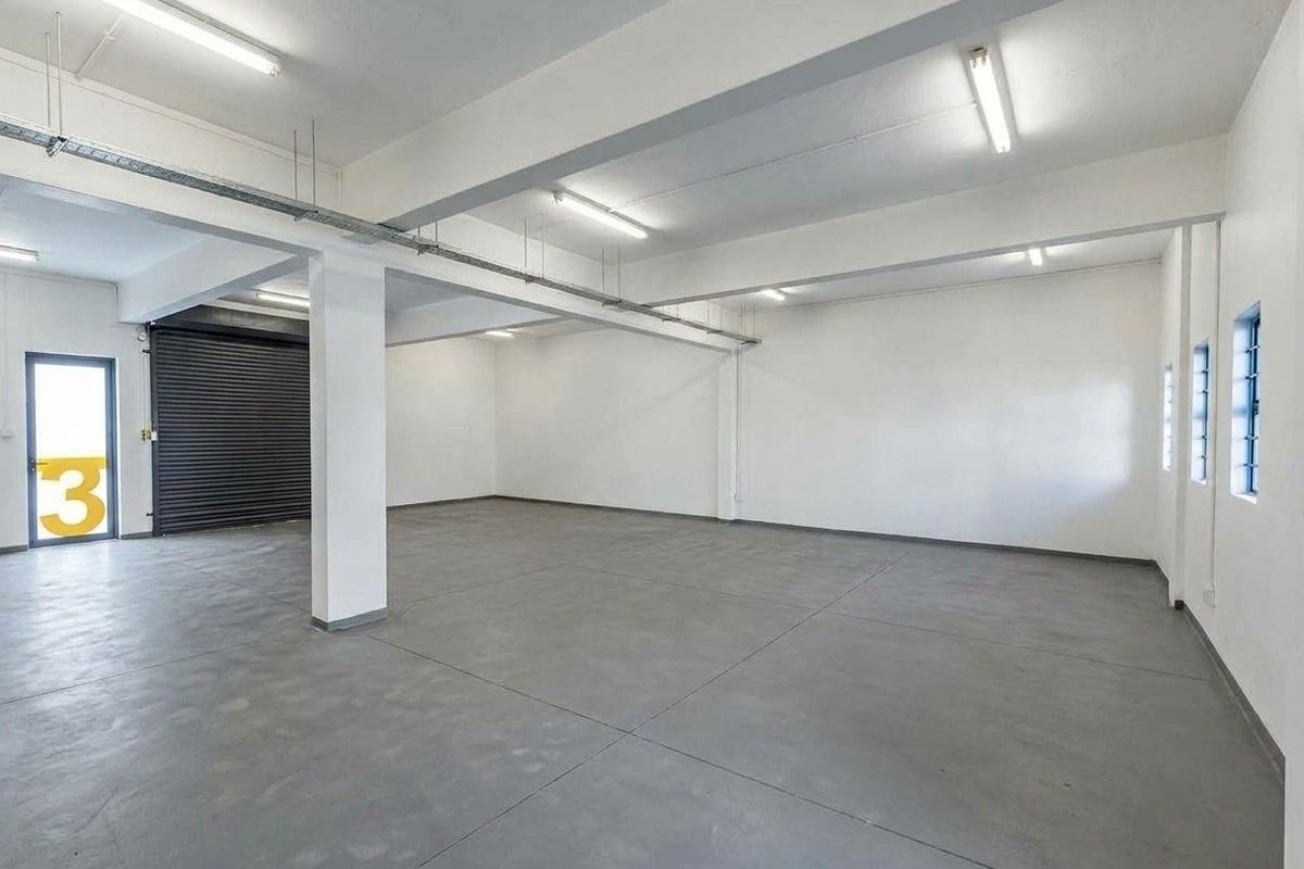 Spacious industrial utility area featuring polished concrete flooring, exposed structural beams, roller shutter door access, and bright overhead fluorescent lighting.