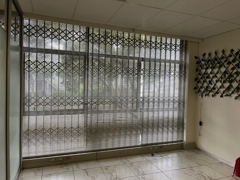 An empty office space featuring large windows with security gates, venetian blinds, tiled flooring, and a suspended ceiling.