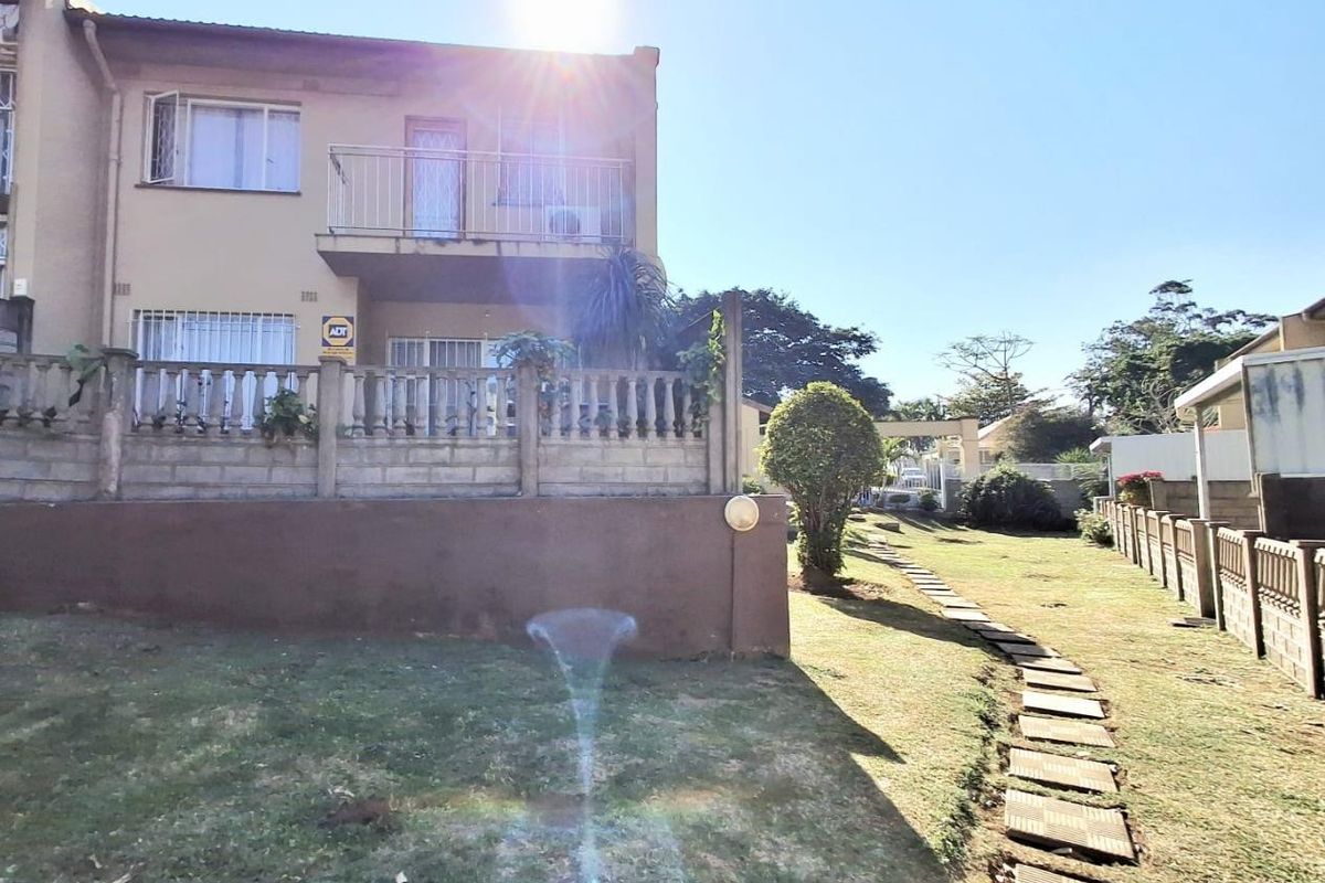 Rear view of the townhouse featuring a private balcony, concrete boundary wall, and a paved garden path leading through the lawn.