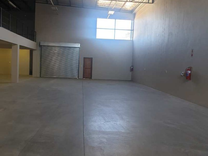 Spacious industrial warehouse unit featuring high ceilings with exposed steel trusses, concrete flooring, a roller shutter door, and a mezzanine level.