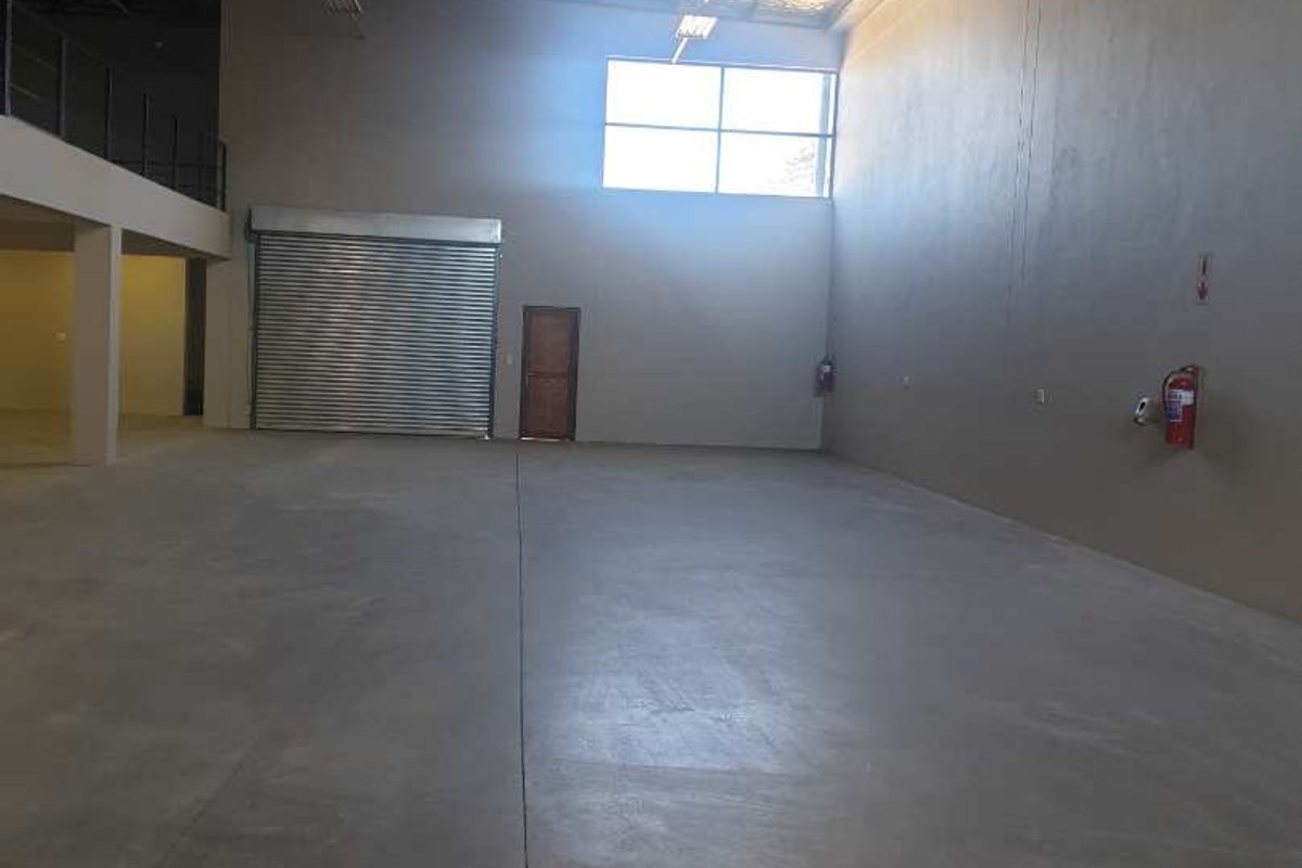 Spacious industrial warehouse unit featuring high ceilings with exposed steel trusses, concrete flooring, a roller shutter door, and a mezzanine level.