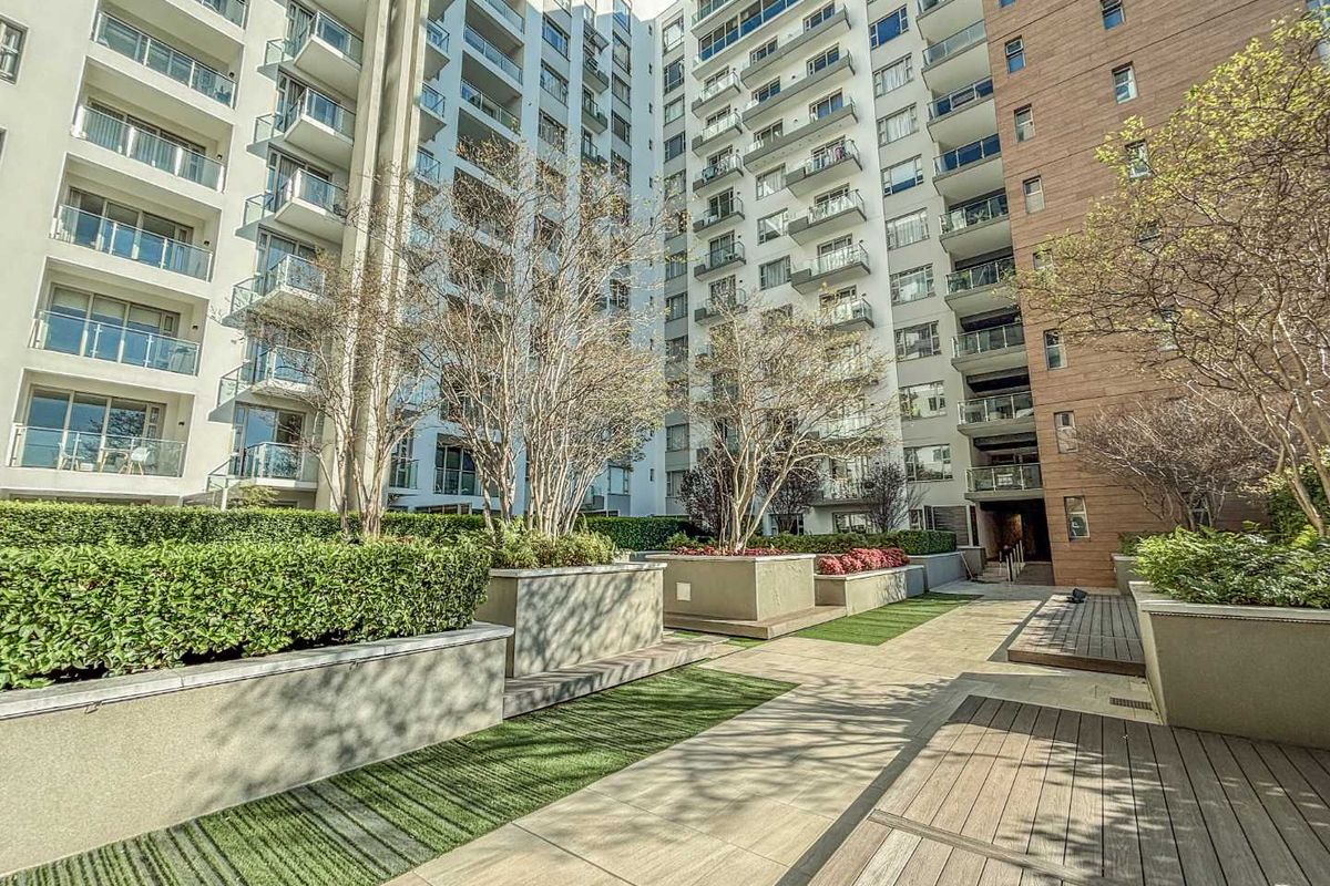 A well-maintained outdoor courtyard area featuring landscaped garden beds, raised planters, and timber decking, situated between modern apartment building facades.
