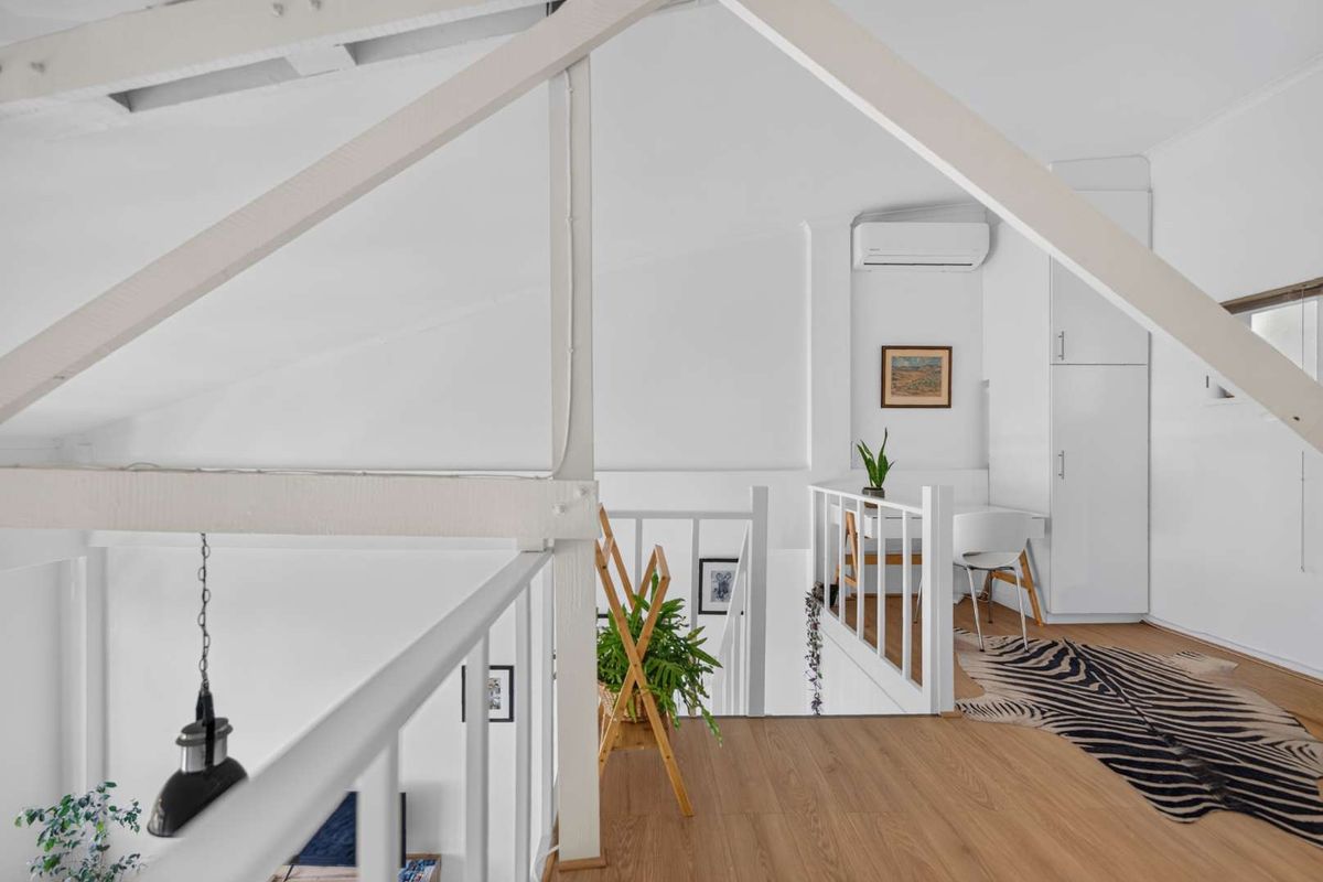 A bright loft-style bedroom featuring exposed timber rafters, light timber flooring, built-in storage, and a wall-mounted split-system air conditioner.