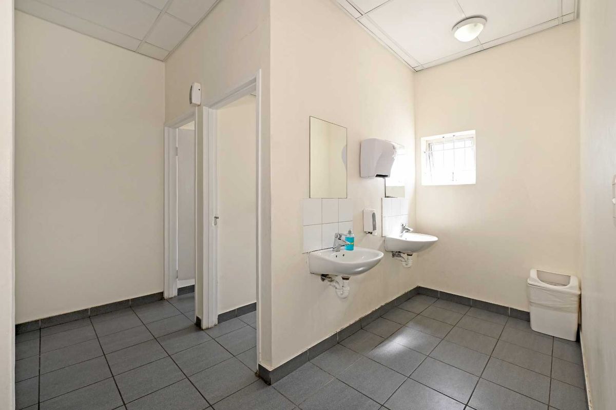 A functional industrial restroom featuring dual wall-mounted ceramic basins, tiled flooring, and a drop ceiling with integrated lighting.