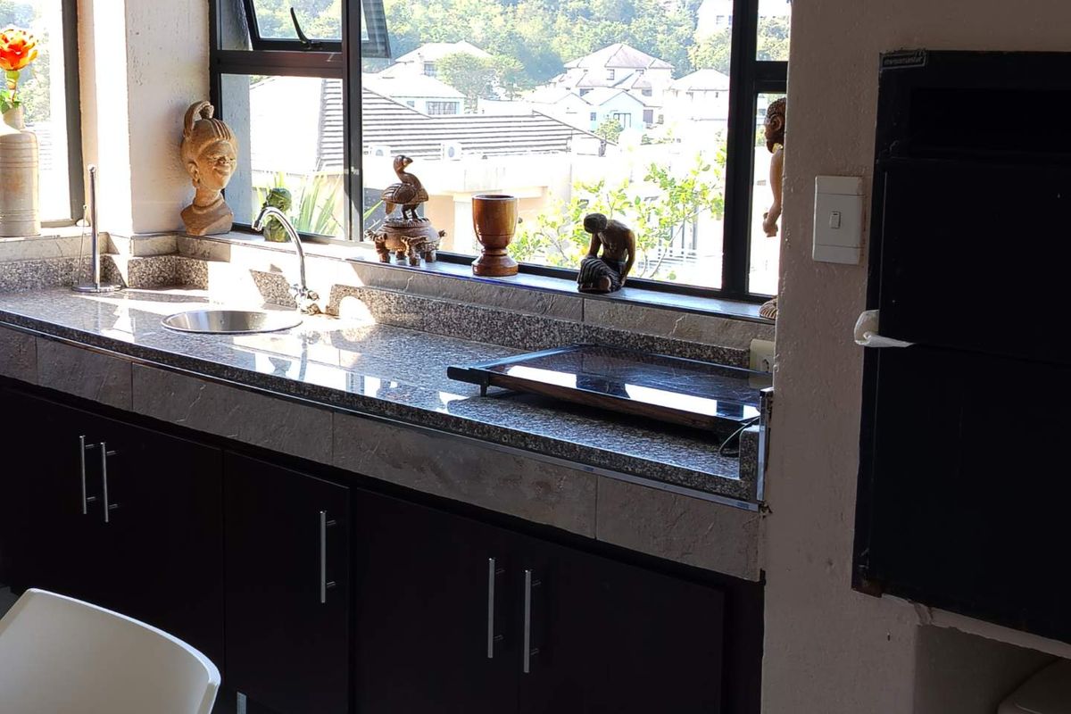A kitchen area featuring dark cabinetry, granite countertops, a stainless steel sink, and large windows providing natural light and neighborhood views.
